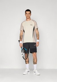 Conjunto deportivo para hombre que incluye una camiseta de manga corta beige y negra y pantalones cortos negros, con una raqueta de tenis y calzado blanco.