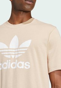 Světle béžové tričko z bavlny, s velkým bílým logem Adidas a třemi bílými pruhy na přední straně. Kulatý výstřih a krátké rukávy.