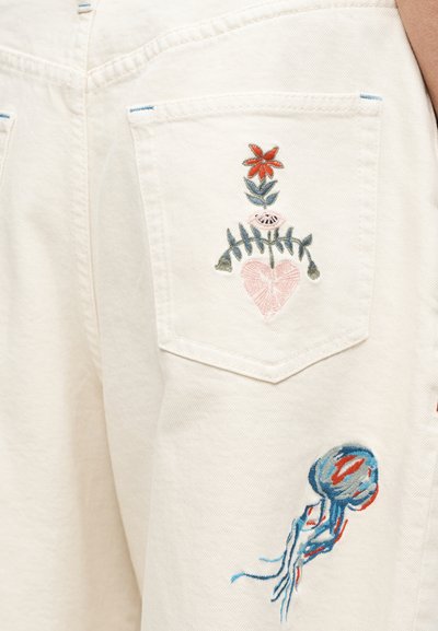 Κρεμ τζιν σορτς με κεντήματα που παρουσιάζουν σχεδιασμούς με λουλούδια και καρδιές, με μοτίβο μέδουσας στις πίσω τσέπες. Υφασμα με υφή και λεπτομερείς ραφές.