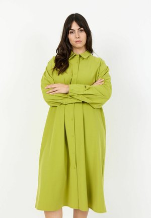 Kobieta o długich ciemnych włosach, ubrana w luźną, zapinaną na guziki limonkowozieloną sukienkę, stojąca z założonymi ramionami na białym tle.