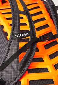 Un sac à dos noir et orange présente un tissu à motifs en grille et un panneau dorsal moulé avec des découpages prononcés pour la ventilation et le soutien.