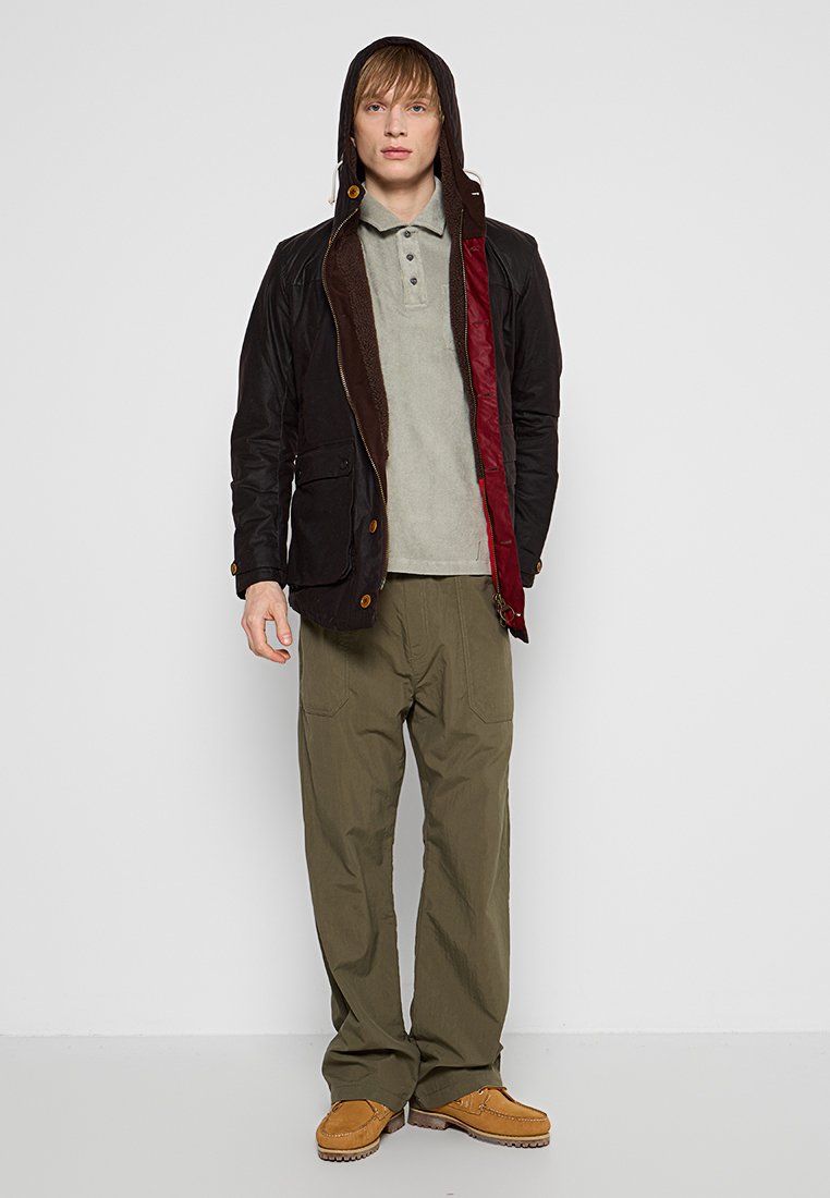 Black and brown hooded jacket with a zip closure, gray polo shirt, olive green pants, and tan shoes, standing on a plain background.