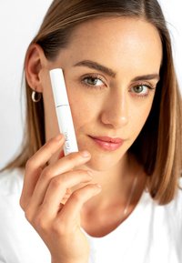 Tubo di mascara bianco con logo in script nero, tenuto da una persona con pelle chiara e capelli castano medio, su uno sfondo neutro.