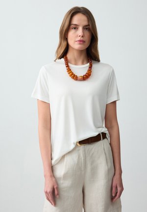 Witte t-shirt van zachte stof met korte mouwen, gedragen met beige linnen broek en een stevige barnstenen ketting in verschillende tinten oranje.