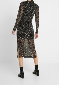 Svart blommig genomskinlig klänning med långa ärmar, figurnära midja och en midjelång kjol med sidoklipp. Bärs med svarta klackade ankelskor.