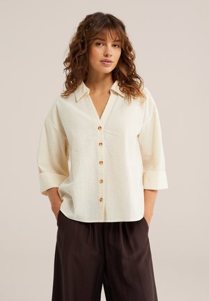 Göndör hajú nő krémszínű, gombos, felhajtott ujjú ingben és sötét, bő nadrágban, kezét zsebre téve, semleges háttér előtt.