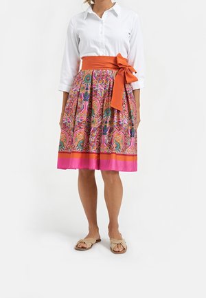 Femme portant une chemise blanche à boutons et une jupe aux motifs colorés atteignant les genoux, avec une large ceinture orange nouée en nœud, sandales beige.