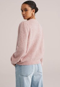 Puha rózsaszín kötött pulóver bordázott mintával, kerek nyakkivágással és lefelé eső vállakkal, világoskék farmerral párosítva.