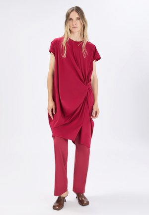 Donna in piedi che indossa una tunica rossa ampia con un nodo laterale, pantaloni rossi abbinati e scarpe con tacco marroni su uno sfondo chiaro uniforme.