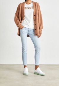 Un cárdigan largo de color marrón claro, una camiseta blanca con letras doradas "BOSS", jeans ajustados de color azul claro y zapatillas blancas con suelas verde menta.