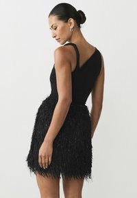 Schwarzes Kleid mit einem eng anliegenden Oberteil und einem gefransten, strukturierten Rock. Ein-Schulter-Design, glattes Material oben, das mit dem fülligen unteren Teil kontrastiert.