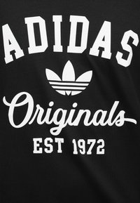 Μαύρο ύφασμα με λευκό λογότυπο Adidas, σύμβολο τριφυλλιού, γραφή "Originals" και το κείμενο "EST 1972".