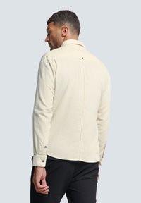 Crème-kleurig langemouwen shirt van zacht materiaal, met een puntkraag, knoopmanchetten en een rugplooi met een enkele zwarte knoopdetail.