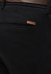 Κοντινό πλάνο στην πίσω τσέπη μαύρων παντελονιών με καφέ δερμάτινη ετικέτα που δείχνει "JACK & JONES" και μέρος από καφέ ζώνη στη μέση.