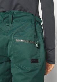 Groene stofbroek met een ritszak, voorzien van een gevoerd ontwerp op de tailleband en een zwart logo-patch. Gevoel van textuur is zichtbaar.