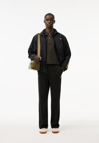 Giacca leggera nera con accenti gialli, polo verde oliva, pantaloni neri e scarpe crema. Tiene una piccola borsa a tracolla verde oliva.