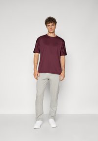 Bordeaux T-shirt met korte mouwen en zwart logo, gecombineerd met lichtgrijze joggingsbroek. Model staat tegen een effen lichte achtergrond.