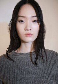 Siv jopič z rebrastim vzorcem in okroglim izrezom, z dolgimi rokavi in subtilno teksturo. Model ima dolge ravne črne lase in minimalen make-up.