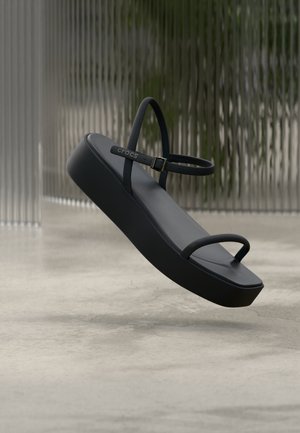 Črni platform sandal z tankimi trakovi in zaponko, blagovne znamke "Crocs", prikazan lebdeč nad gladkim tlakom pred ozadjem iz rebrastega stekla.