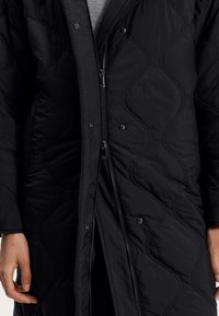 Veste matelassée noire avec une fermeture éclair à l'avant et des boutons-pression. Le tissu est lisse avec une légère brillance et un motif en vagues texturé.