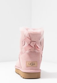 Bottine en daim rose avec doublure douce en fausse fourrure, nœud ruban à l'arrière, et semelle extérieure en caoutchouc. Logo UGG sur le talon.