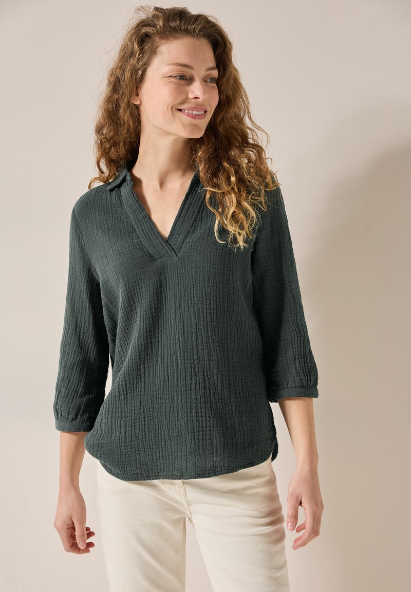 Blusa verde scuro, strutturata, con scollatura a V e maniche a tre quarti, abbinata a pantaloni chiari. Leggero motivo di pieghe visibile.