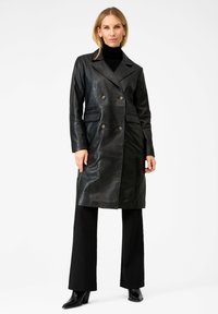 Vrouw die staat in een zwart dubbele leren jas, zwarte coltrui, zwarte broek en zwarte hakken tegen een effen achtergrond.
