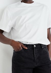 Camiseta de algodón blanca con cuello redondo, combinada con jeans de mezclilla negros que tienen un bolsillo frontal y un detalle de botón plateado.