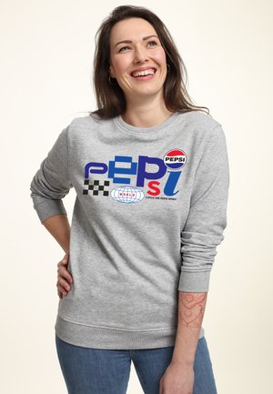 Felpa grigia con grafiche tra cui "PEPSI" e un motivo a scacchi. Polsi e orlo a coste, vestibilità morbida. Jeans con una sottile texture.