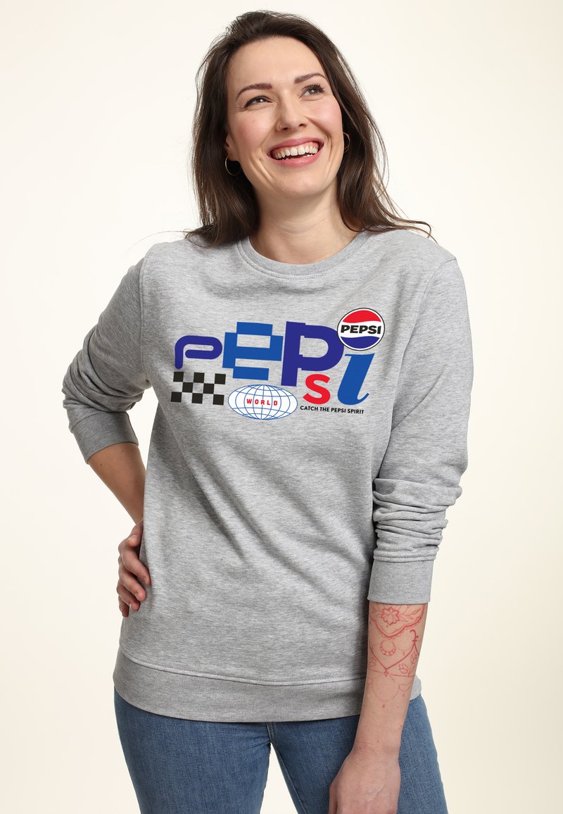 Felpa grigia con grafiche tra cui "PEPSI" e un motivo a scacchi. Polsi e orlo a coste, vestibilità morbida. Jeans con una sottile texture.