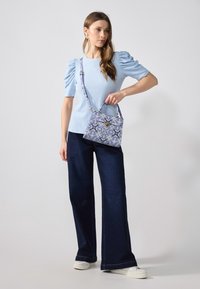 Haut en maille bleu avec manches bouffantes, associé à un jean large foncé et des baskets blanches. Le sac présente un motif floral en bleu et or.
