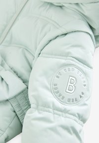 Svijetlozelena puffer jakna s quilted teksturom i elastičnim manžetama. Sadrži kružni pečat s uzdignutim logom "B" na rukavu.