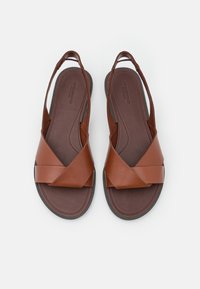 Sandales en cuir marron avec un design à lanière croisée, semelle plate et semelle intérieure texturée. Le style à dos nu expose le talon.