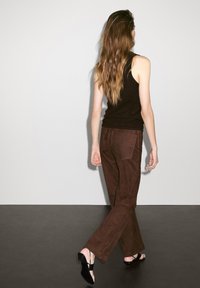 Pantaloni scamosciati marroni con un design a gamba dritta, abbinati a tacchi neri con cinturini. La parte superiore è un top nero aderente. Il modello è mostrato di spalle.