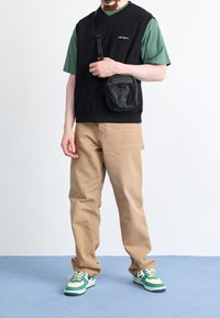 Felpa nera senza maniche sopra una maglietta verde, pantaloni beige e scarpe da ginnastica verdi. Piccola borsa a tracolla nera con cerniere e dettagli in metallo.