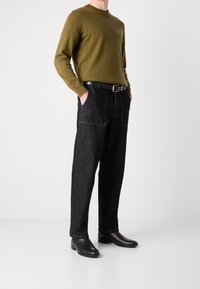 Chemise à manches longues vert olive, pantalon en jean noir avec coutures visibles, et bottines en cuir noir avec une finition texturée.