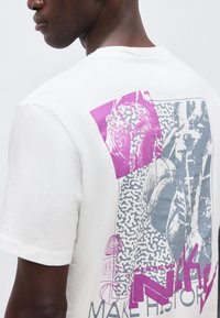 Άνδρας φοράει λευκό μπλουζάκι με αφηρημένο μοβ και γκρι γραφικό σχέδιο και μερικό κείμενο στην πλάτη.