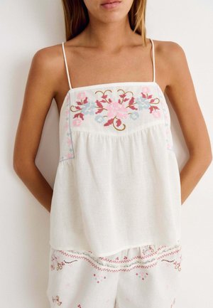 Femme portant un haut blanc sans manches avec broderie florale rose et bleue et un pantalon ample assorti avec une broderie similaire.