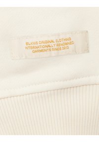 Witte stoffen label met gouden tekst, met de tekst "BLKWS ORIGINAL CLOTHING" en "GARMENTS SINCE 2013." Katoenen textuur met geribbelde rand.