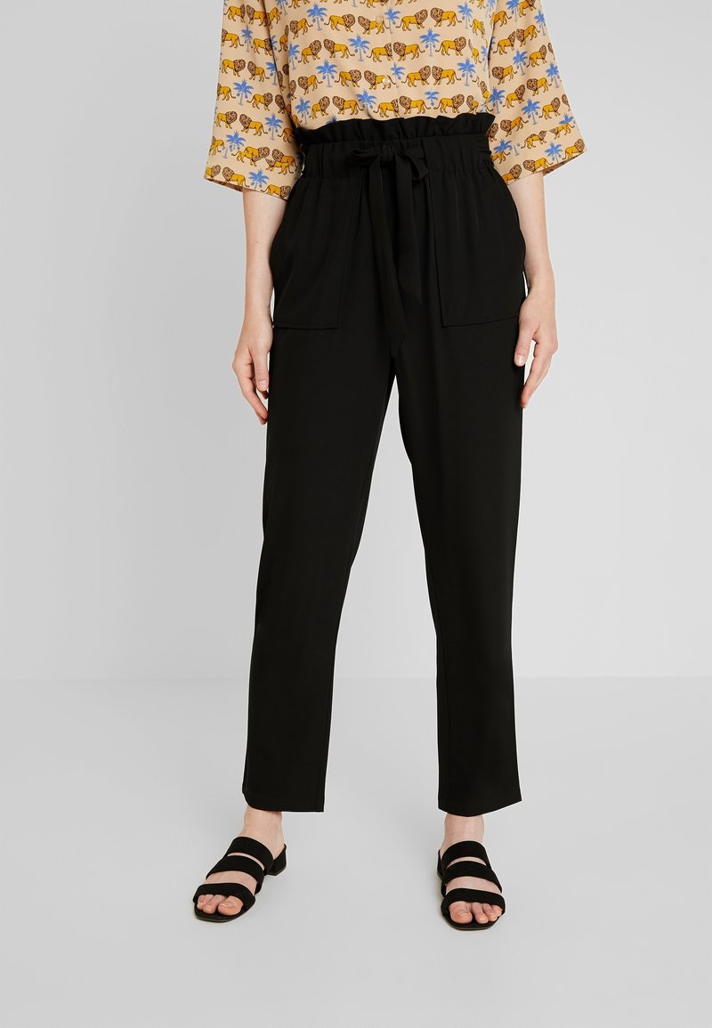 Pantalon noir taille haute avec taille paperbag et ceinture à nouer, porté avec des sandales noires à double bride et un chemisier à manches courtes à motif.