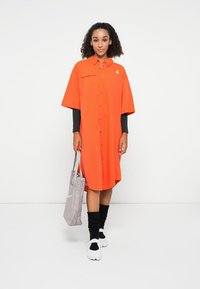 Abito camicia arancione oversize con maniche corte, una tasca frontale, abbinato a una borsa a tracolla grigia e calzini neri. Scarpe bianche completano il look.