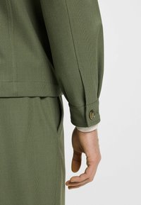 Olivgrön jacka med texturerad tyg, synlig sömnad och detaljer med knäppning vid ärmen. Handen placerad vid sidan.