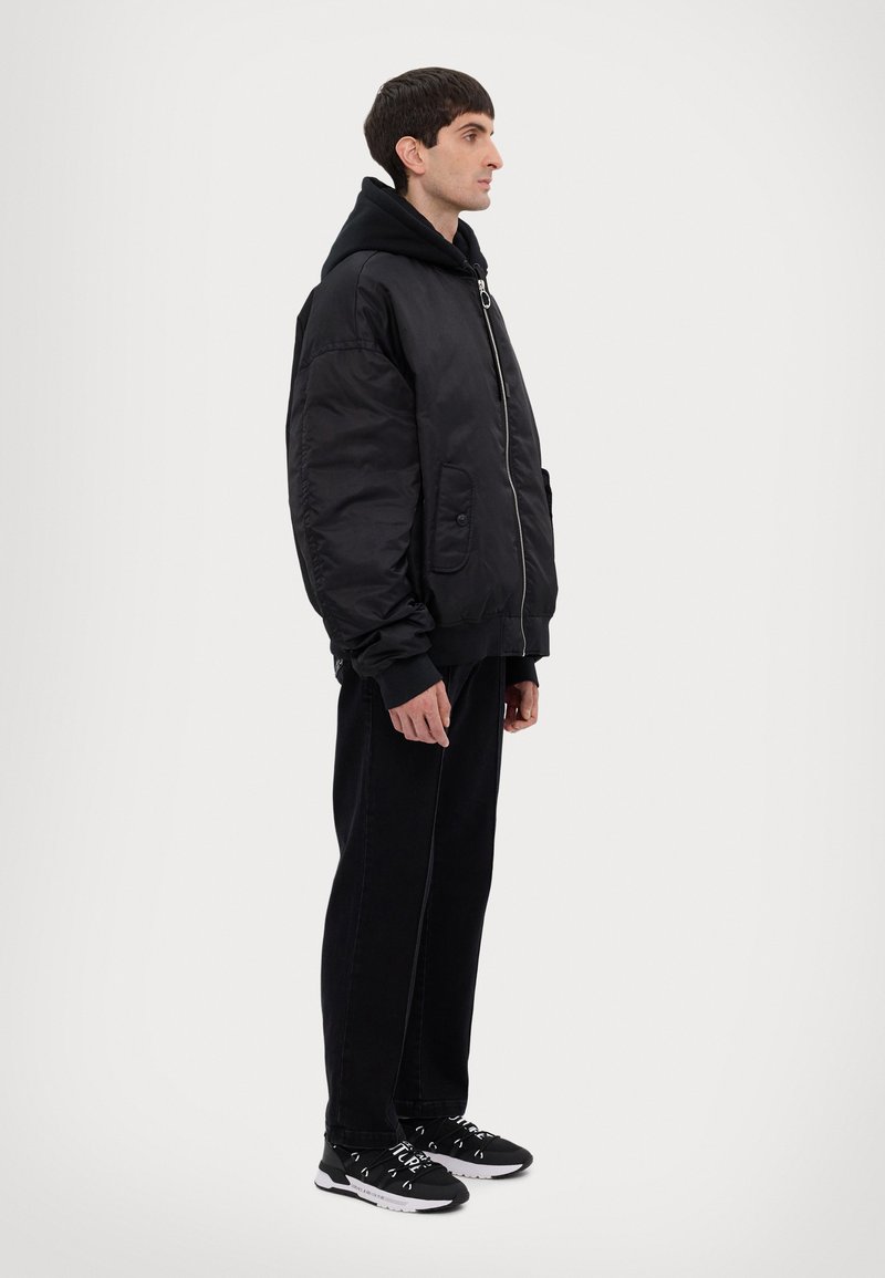 Giacca bomber nera con cappuccio, chiusura con zip e due tasche frontali. Indossata con pantaloni neri e scarpe da ginnastica nere. Design semplice.