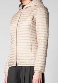 Veste matelassée beige clair avec une coupe ajustée, texture matelassée horizontale, fermeture éclair à l'avant et capuche. Tissu doux avec manches froncées.