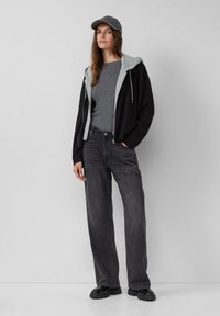 Vrouw in een grijze pet, grijs geribbeld lang shirt, zwarte hoodie met ritssluiting en grijze voering, zwarte wijde jeans en zwarte chunky schoenen.