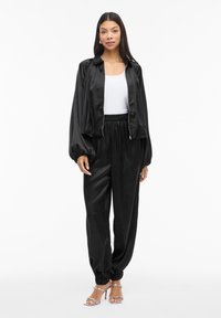 Krótka czarna satynowa kurtka z elastycznymi mankietami, w komplecie z pasującymi, wysokim stanem spodniami z elastycznym wykończeniem nogawek. Całość uzupełniają sandały na przezroczystym obcasie.