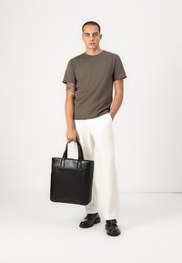 Schwarze Leder-Umhängetasche mit strukturiertem Design und zwei stabilen Griffen, kombiniert mit einem braunen T-Shirt und weißen Hosen. Glatte Textur.