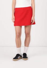 Fustă mini roșie dintr-un material neted, având un design simplu. Asortată cu adidași negri și șosete albe, pe un fundal simplu.
