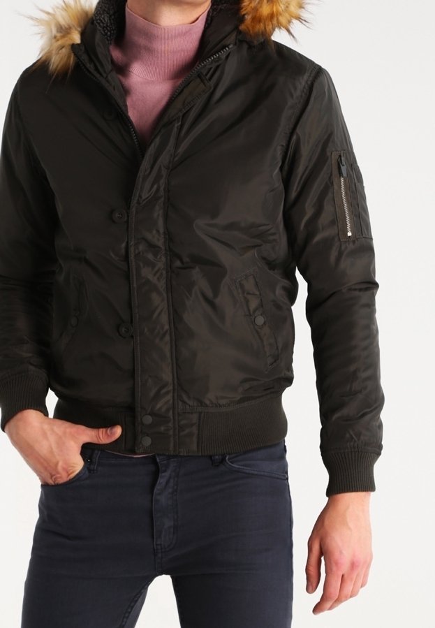 Homme portant une veste bomber noire à capuche bordée de fourrure et poignets côtelés sur un pull à col roulé rose, associé à un jean sombre.