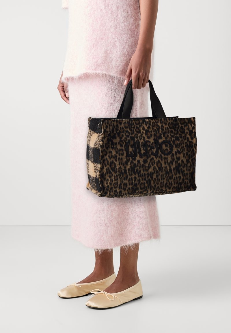 Torebka w zwierzęcy wzór panterki z czarnymi akcentami, z krótkimi uchwytami i logo, trzymana przez osobę w miękkim, różowym, futrzanym ubraniu i beżowych balerinach.
